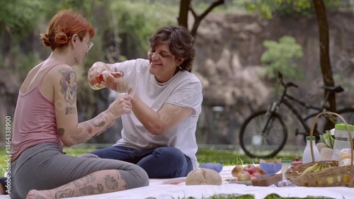 Happy LGBTQ+ couple enjoying a romantic picnic in nature, opening a bottle of rose wine near a scenic waterfall.