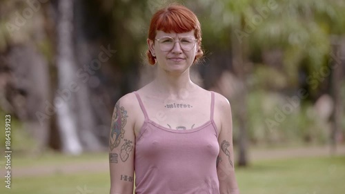 Portrait of a confident young woman with red hair and tattoos standing in a park.