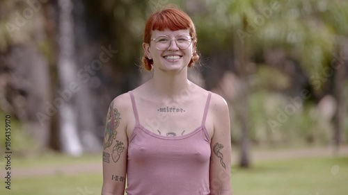 Portrait of a smiling young woman with red hair, tattoos, and glasses in a lush park setting.