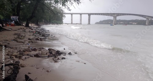 Rainy day view of the sandy and rocky shore at a beach with waves and Techo Morakat Bridge in Sihanoukville.