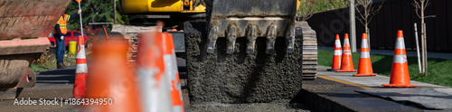 Excavator bucket attachment removing section of asphalt cut in road to dig a utilities trench, public infrastructure construction site, workman watching the project
