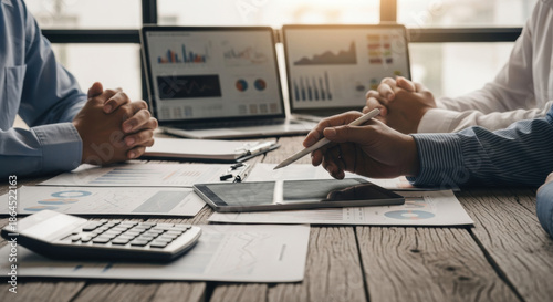 A close up shot showcases a business team reviewing financial data on a wooden table, with laptops, documents, and a digital tablet creating a workspace scene.