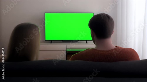 Couple enjoys evening entertainment, watching a television with a bright green screen, perfect for creative content replacement