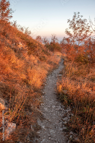 The mountain trail climbs up the slope, suggesting a hiking route. The trail is rocky and surrounded by dry grass and autumn vegetation at sunset. The warm lighting, with orange and red hues, creates 