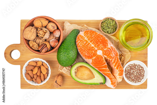 Top view of healthy fats food sources including raw salmon steak, avocado, nuts, seeds and olive oil arranged on wooden cutting board, balanced nutrition and clean eating concept