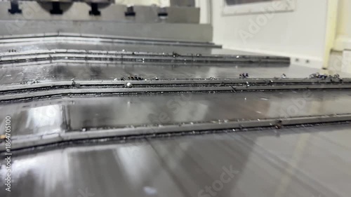 Milling machine worktable in action: sturdy metal surface with T-slots grips workpiece firmly as spindle spins above. Chips scatter, coolant flows, precise feeds along X-Y axes. 