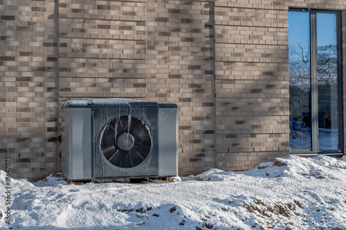 Air source heat pump unit with icicles hanging from casing installed against modern brick building surrounded by snow  during winter. Energy-efficient residential heating technology.