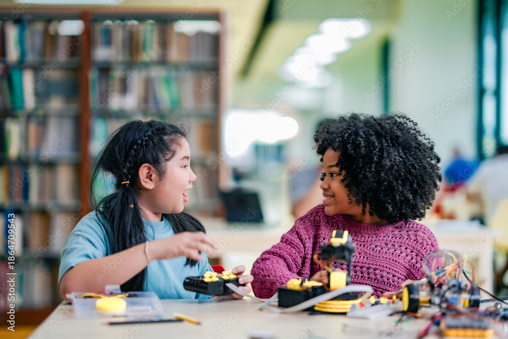 Fototapeta premium African American girl guides robotics experiment alongside Asian girl, problem solving activity. Black girl develops technology skills through shared robotics work, classroom collaboration.