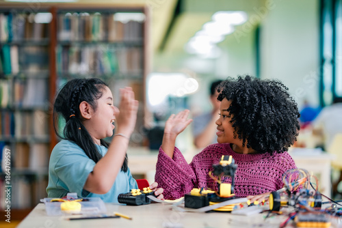 Tablou pe pânză Two children celebrate robotics project success inside library, smiling with raised hands, robotic arm kit, teamwork learning, STEM education, creativity, confidence, joyful study moment