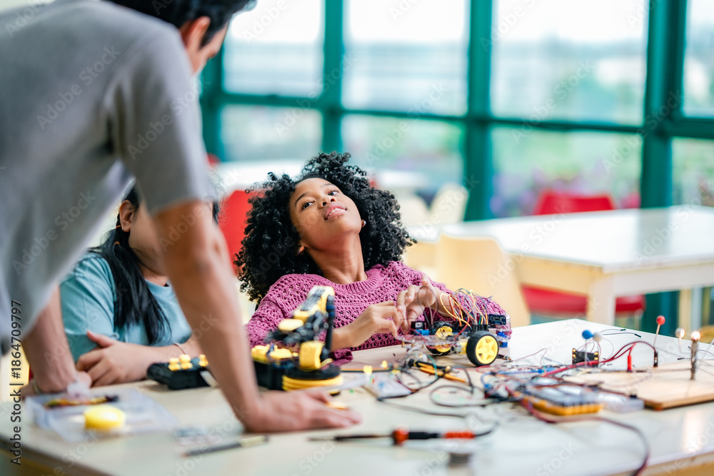 Fototapeta premium Caucasian teacher supports diversity girl during STEM robotics lesson, guiding electronics practice, encouraging diversity learning, technology skills development, collaborative education environment.