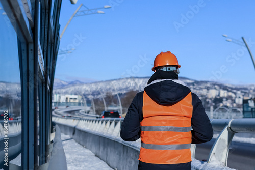 Rear view of worker in orange vest surveying winter highway bridge