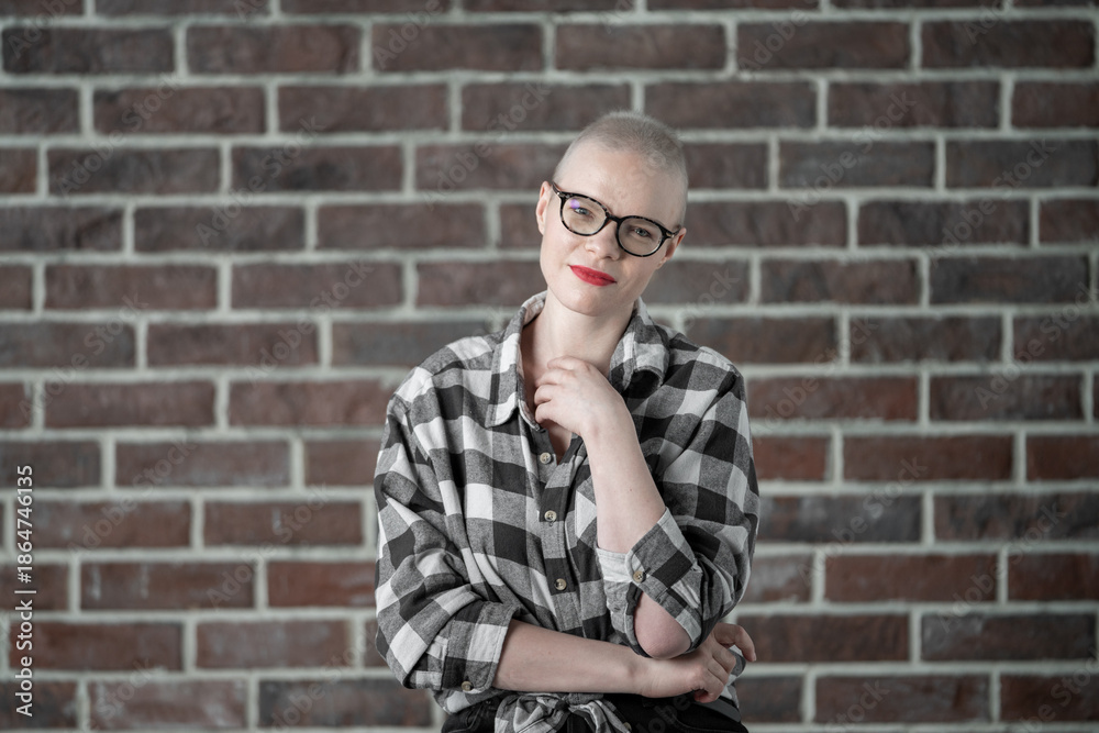 Obraz premium Portrait of a beautiful young caucasian woman with shaved head. Female cancer survivor. Smiling Woman with Glasses Posing Confidently in a Studio Environment. Model Has Glaucoma. Waering Plaid Shirt