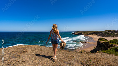 Beautiful middle-aged woman tourist walking on cliff overlooking Monte Clerigo beach and ocean in summertime. Algarve Portugal. Back view	