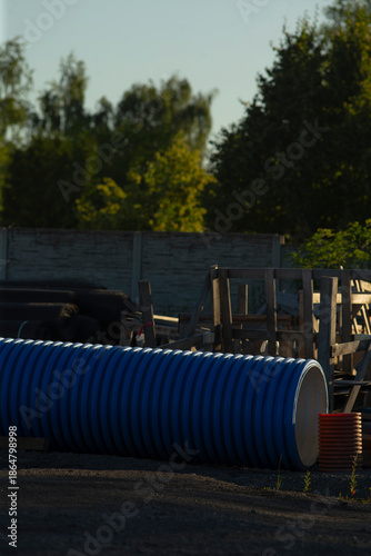 blue pipe stacked in industrial yard under warm dusk light, corrugated surface catching soft highlights, scattered pallets and fencing framing background, tall trees and concrete wall