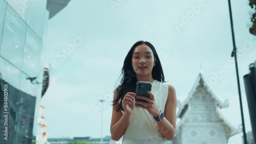 Happy young asian woman walking in the city and looking at her smartphone with a smile