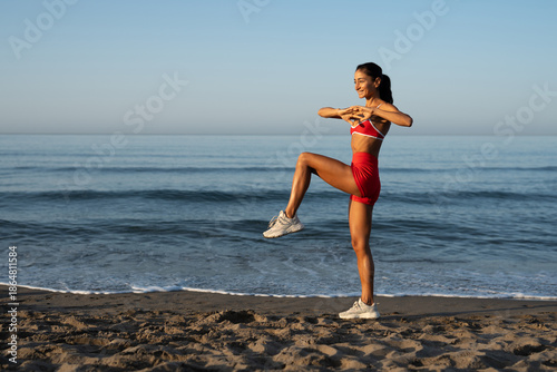 Fit young woman performing balance training with one knee lifted on a sandy beach at sunrise, wearing red athletic outfit, enjoying outdoor morning workout with ocean waves in the background