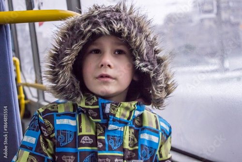 Little child looking through the window: a boy in a bright winter jacket with a faux fur hood and gloves. 