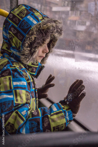 Little child looking through the window: a boy in a bright winter jacket with a faux fur hood and gloves. 