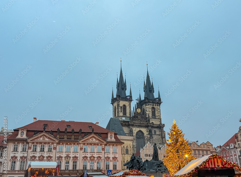 Fototapeta premium The Gothic twin towers of the Church of Our Lady before Týn rise behind a bright Christmas tree and the Jan Hus Memorial in Old Town Square, capturing a festive winter atmosphere in Prague, Czechia.