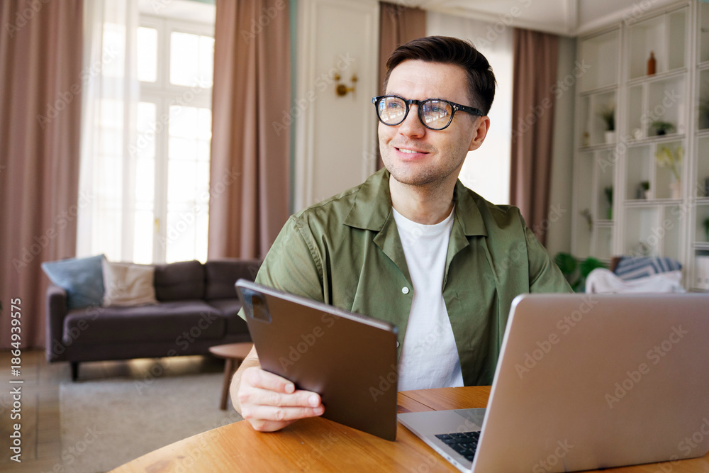 Fototapeta premium Young man sits at table using tablet and laptop
