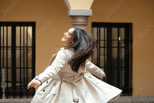 Quadro em tela Young woman with long dark hair wearing a light trench coat joyfully spins outdo