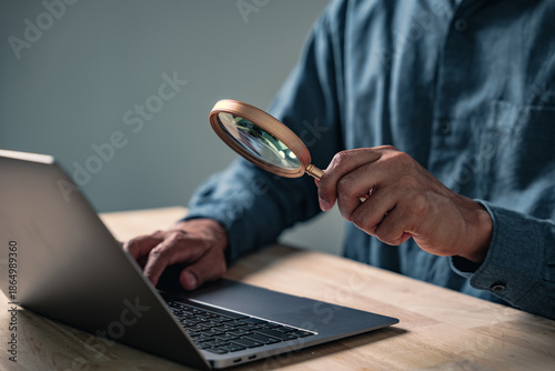 Obraz na plátne A professional man makes a hand gesture in an investigative business pose, closely examining a computer while posing with a magnifying glass for data research and analysis concepts