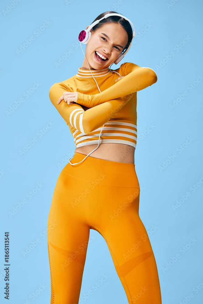 Naklejka premium Smiling young woman in bright yellow activewear and headphones poses energetically against a blue studio background, showcasing fitness, sport, movement and joyful workout lifestyle.