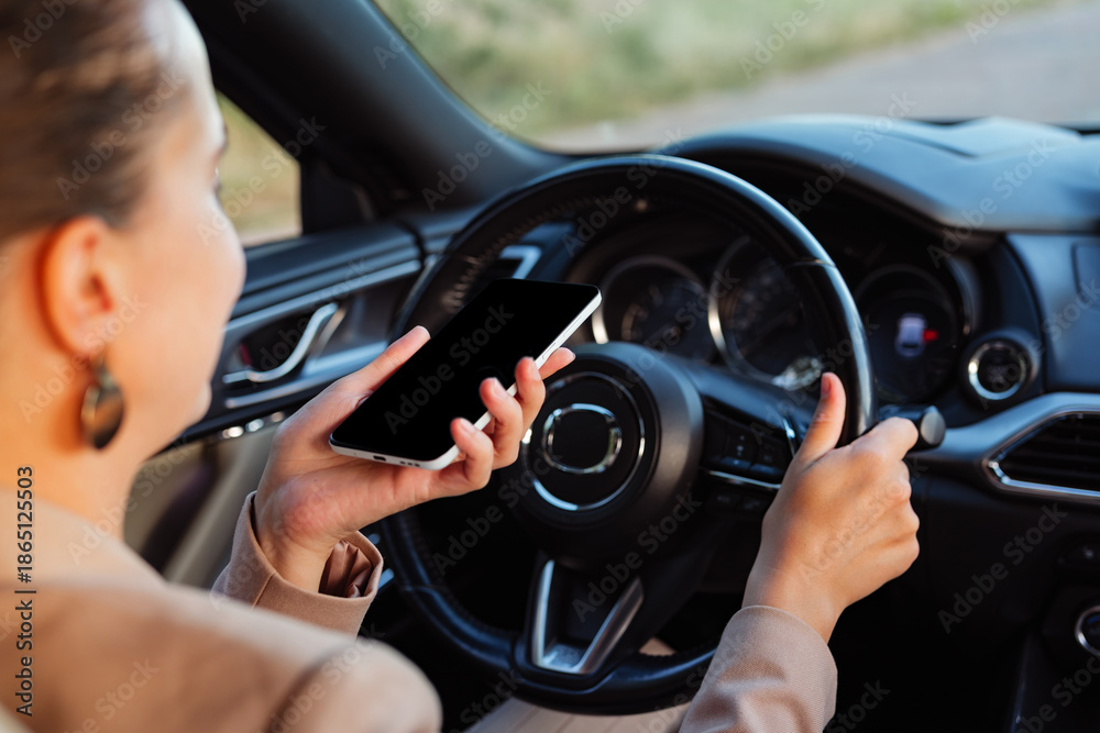 Naklejka premium A woman sits in the driver's seat of a car, holding a smartphone in one hand while steering with the other. The scene is set on a road under clear skies