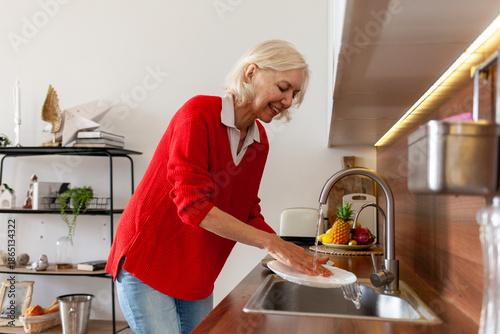 Photography elderly woman in red blouse washes dishes at home in the kitchen, gray-haired gr
