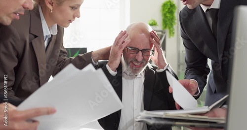 Office businessman shows stress during deadline pressure. Colleagues crowd his paperwork as the team demands as he holds his head, headache and anxiety rising. Strong concept of office burnout.