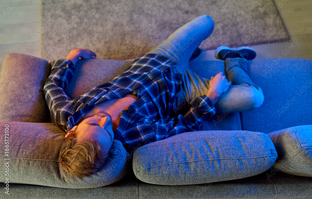 Fototapeta premium Man with drug or alcohol addiction lying on sofa at home in depression, top view. Lonely male addict suffering from hangover after booze party or overdose, poisoning by toxic narcotic substance