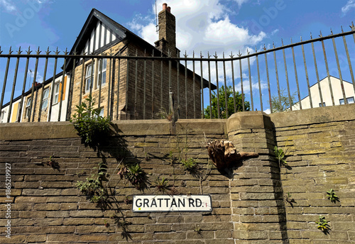 A Victorian stone wall carrying the Grattan Rd sign is partly claimed by overgrown plants. Beyond the fence, a steep roofed house and chimney rise into a clear, vibrant sky in Bradford, Yorkshire, UK