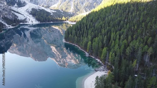 Forward drone flight over Lake Braies approaching dense pine forest along the turquoise shoreline. Alpine mountain landscape in the Dolomites, Italy.