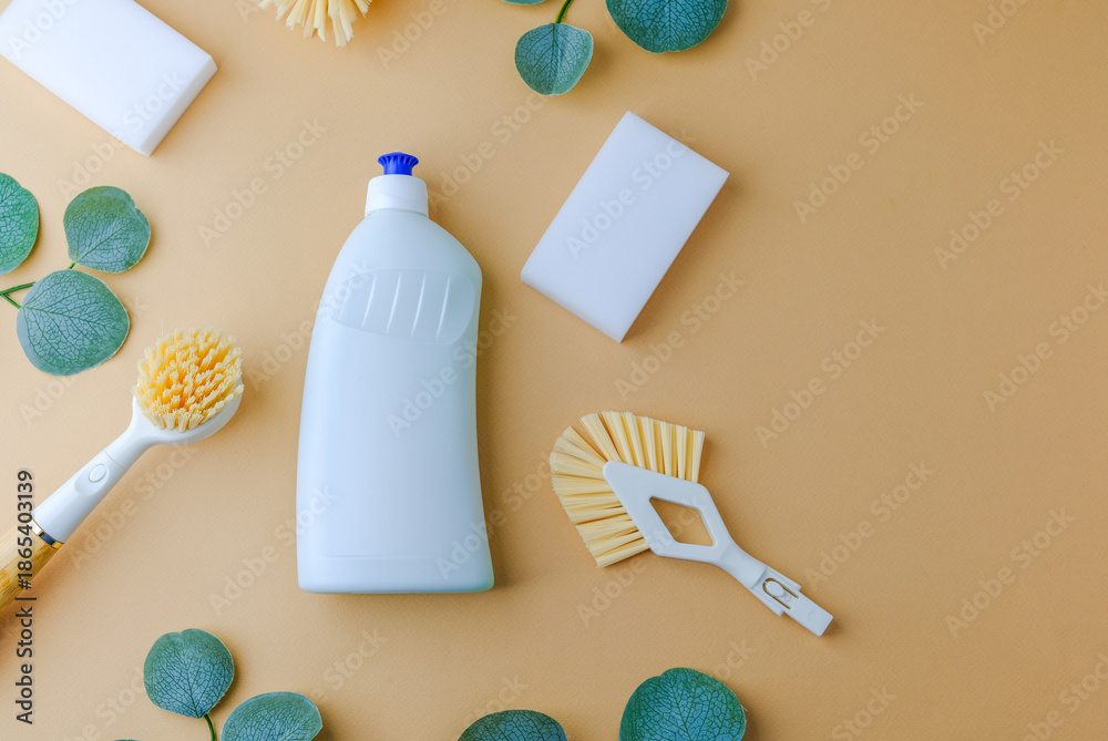 Obraz premium White bottle with cleaning brushes and sponges, natural green leaves, simple home cleaning supplies on light brown background.