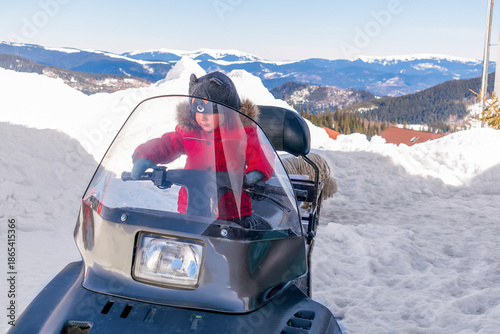 Little kid child boy sitting on modern snowmobile scooter vehicle at winter holiday,ski alpine resort in mountains outdoor nature landscape,Europe.extreme sport activities.adventure trip