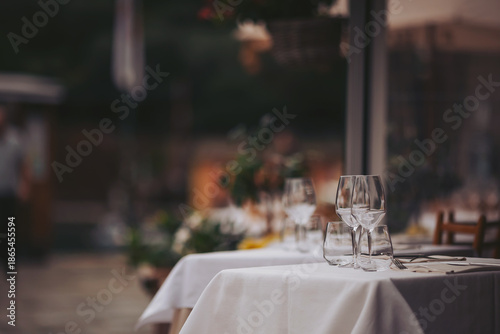 A dining table is set with several glasses placed on a clean tablecloth. The restaurant is located outdoors and shows some blurred background elements in the evening light