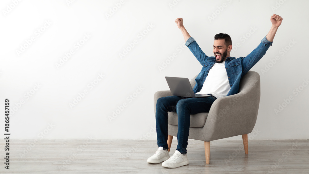 © Prostock-studio - Overjoyed millennial Arab man sitting in armchair with laptop, celebrating online win or work success against white studio wall, copy space. Young Eastern guy enjoying big sale in web store, banner © Prostock-studio - Overjoyed millennial Arab man sitting in armchair with laptop, celebrating online win or work success against white studio wall, copy space. Young Eastern guy enjoying big sale in web store, banner