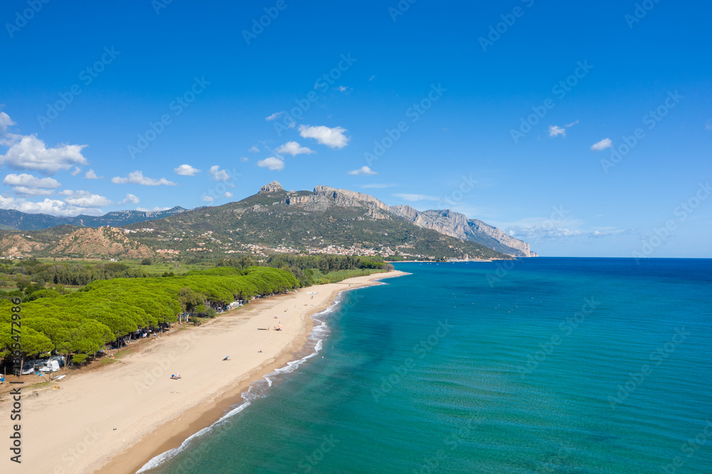 Fototapeta premium Aerial view of a tranquil sandy beach bordered by lush pine trees and clear blue sea, with dramatic mountains rising in the background near Lotzorai, Sardinia. Bright sunlight and a vivid sky enhance