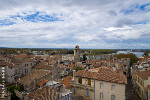 Konstfotografi Wide view over the terracotta rooftops of Arles with a historic stone tower and the Rhone River winding through the landscape beneath a dramatic, cloud-filled sky