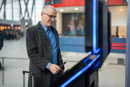 Foto Mature man using smartphone at airport check-in kiosk