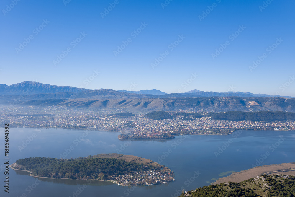 Obraz premium Elevated perspective of the tree-covered island of Ioannina set in calm lake waters, with the city of Ioannina and the rugged Pindus mountains forming a dramatic backdrop in Epirus, Greece. Crisp