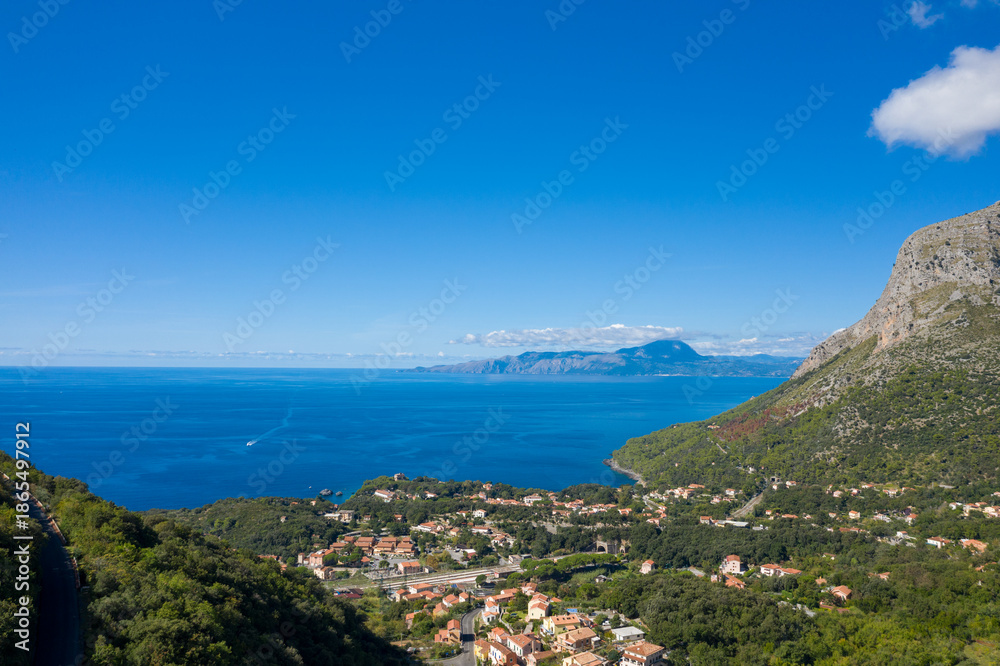 Obraz premium Wide-angle scene showing Maratea village nestled among green hills overlooking the deep blue Tyrrhenian Sea, with rugged mountains in the distance under a clear, sunny sky. The vivid colors and open