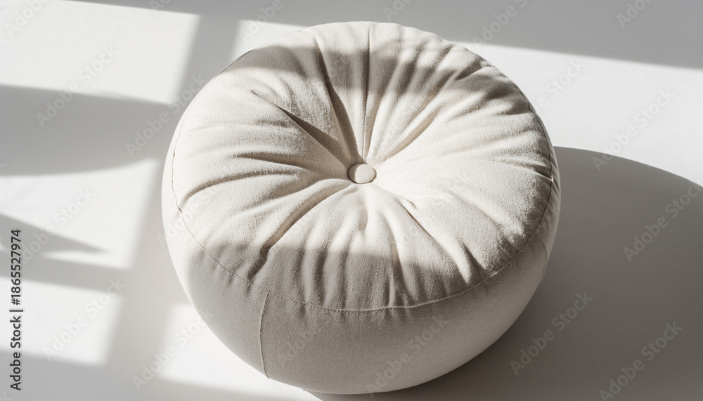 Obraz premium Minimalist round off-white floor cushion or pillow with tufted button detail, casting long shadows on a bright white background, showcasing texture and soft light.