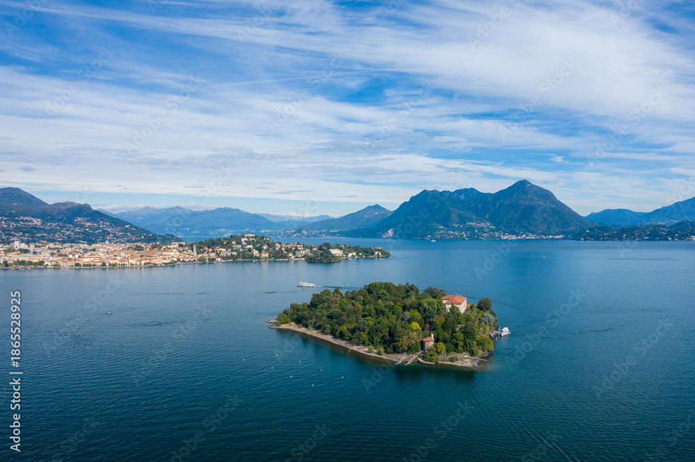 Obraz premium A wide aerial perspective captures Isola Madre surrounded by the deep blue waters of Lake Maggiore, with distant towns, forested hills, and rugged alpine mountains under a sky streaked with soft