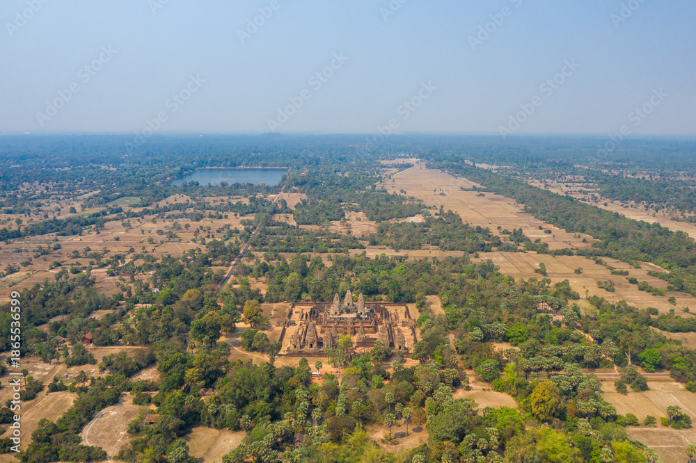 Obraz premium Expansive aerial perspective of the ancient Pre Rup temple complex surrounded by dry fields, scattered trees, and distant water reservoirs in the Angkor region of Cambodia. The scene captures the