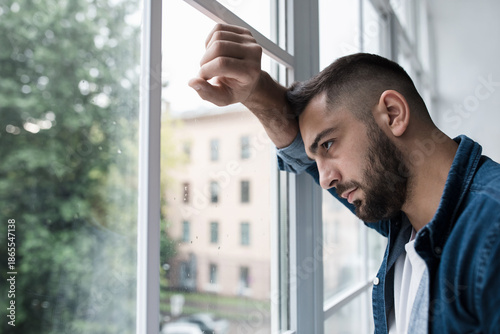 Dramatic portrait of caucasian man, alone by window at home looking sorrow with desperate feeling and depressed from unemployment. Sad unhappy young handsome man with hand on glass in kitchen interior