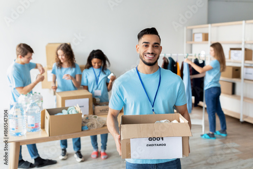 Canvas Print Young arab male volunteer in blue uniform joyfully holds a cardboard box labeled food donations