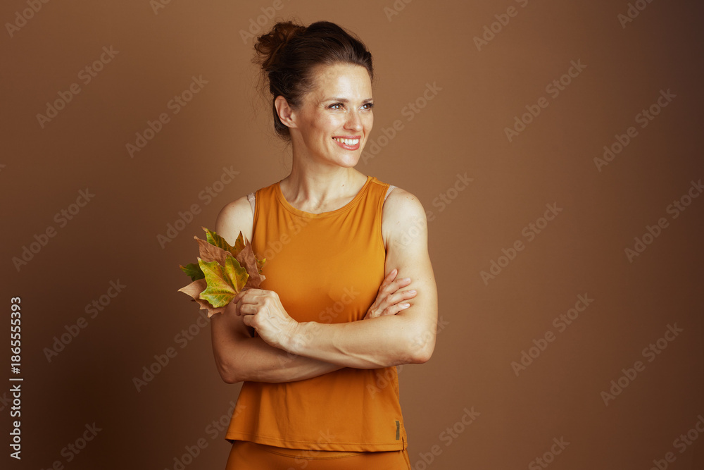 © Alliance - A smiling middle-aged woman in a mustard yellow tank top stands with crossed arms, holding autumn leaves, looking right with a content expression, conveying autumnal serenity.