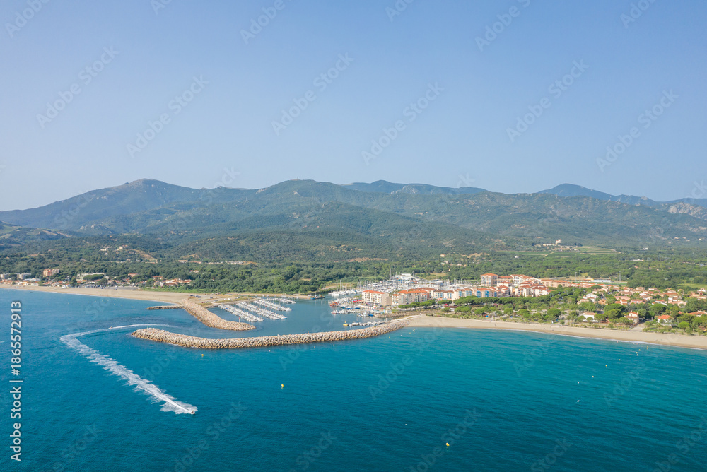Fototapeta premium Aerial view of a marina with boats docked along a curved breakwater, bordered by turquoise water and a sandy beach, with lush green hills and mountains rising in the background under a clear blue sky.