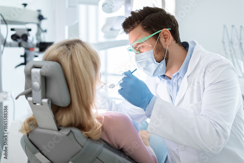Obraz na płótnie Man dentist in face mask and glasses doing treatment for patient blonde lady, holding dental tools, wearing rubber gloves